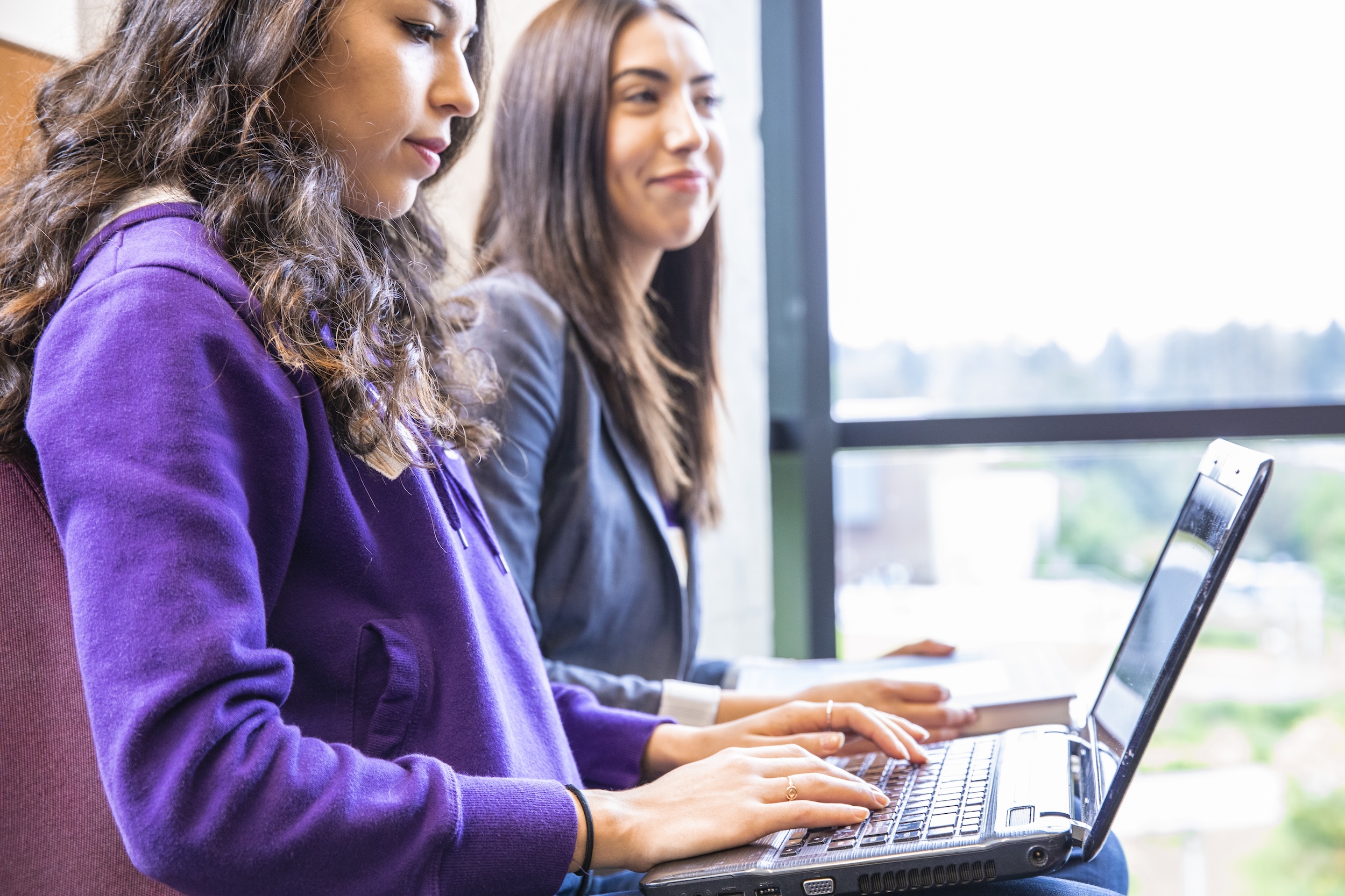 two students, one looking forward, one looking at and working on her laptop