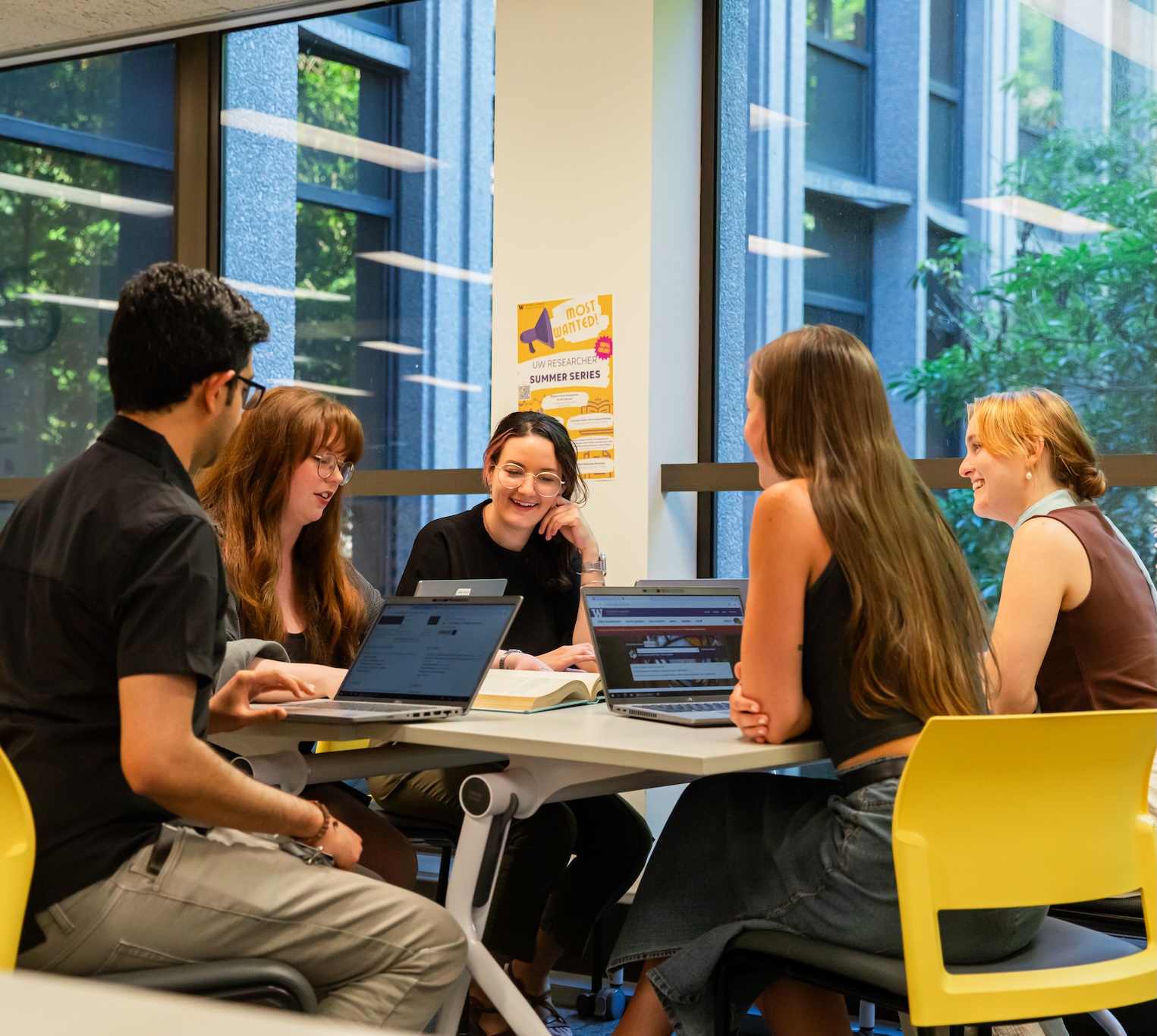A group of 5 students sitting at a table with their laptops open, chatting and smiling